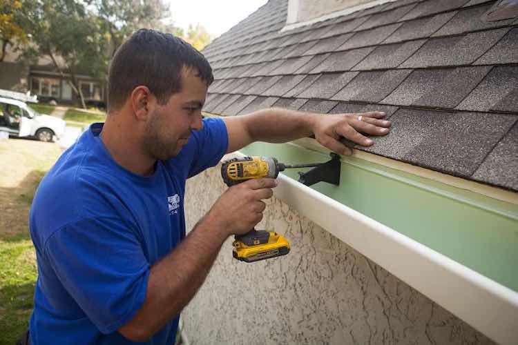 Man in Blue Shirt Putting New Gutters on Home Using Drill - K-Guard St. Louis
